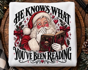 May include: A white sweatshirt featuring a graphic of Santa Claus with a white beard and red hat, holding an open book with a skull and rose design. The text reads HE KNOWS WHAT YOU'VE BEEN READING, surrounded by red bows, holly, and hearts.