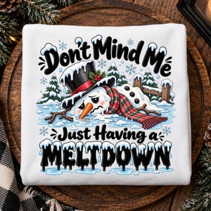 May include: A white sweatshirt featuring a graphic of a melting snowman wearing a black top hat with a red band and a red and green plaid scarf. The text reads "Don't Mind Me Just Having a MELTDOWN" in black, with snowy pine trees and snowflakes.