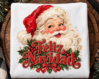 May include: A white sweatshirt featuring a graphic of Santa Claus with a red hat and white beard, positioned above the red text Feliz Navidad, surrounded by green holly leaves, red berries, and a red ribbon bow at the bottom.