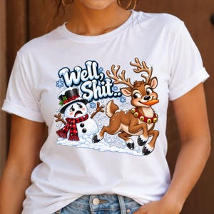 May include: A white t-shirt featuring a graphic print of a distressed snowman wearing a black top hat and a red and black plaid scarf, alongside a brown reindeer with a bell collar holding a carrot, with the text Well, Shit above them.