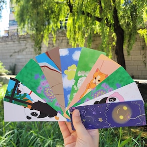 May include: A collection of rectangular bookmarks fanned out, featuring various colorful illustrations including a panda, a cat, a blue sky with clouds, and a dark purple night scene with stars and a glowing yellow moon-like shape.