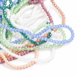 May include: A collection of multi-colored faceted glass beads arranged in loose, overlapping strands. The assortment includes translucent white, light blue, emerald green, pale pink, peach, and copper-toned beads with a reflective finish.