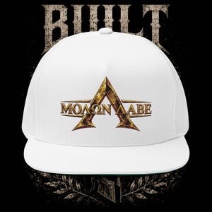 May include: A white baseball cap featuring a large, distressed gold-toned letter A with the Greek text "ΜΟΛΩΝ ΛΑΒΕ" centred across it. The cap has a structured crown and a flat brim, with a dark, weathered graphic design visible behind the cap.