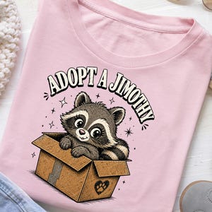 Könnte beinhalten: Ein hellrosa T-Shirt mit einer grafischen Illustration eines grau-weißen Waschbären, der in einem braunen Pappkarton sitzt. Der Text ADOPT A JIMOTHY ist in einer schwarzen, geschwungenen Schrift über dem Waschbären gedruckt, umgeben von kleinen Sternen.