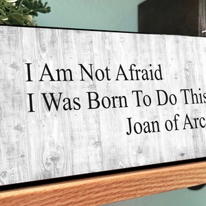 May include: A rectangular wooden sign with the quote "I Am Not Afraid I Was Born To Do This Joan of Arc" in black text. The sign has a gray wood grain pattern and is displayed on a wooden shelf.