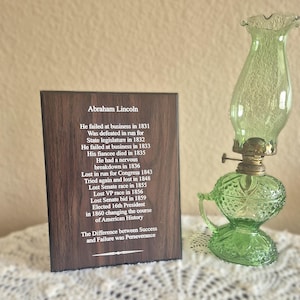 May include: A rectangular dark wood plaque featuring white text detailing the life events of Abraham Lincoln, positioned next to a vintage-style green glass oil lamp with a textured base and a clear chimney top.