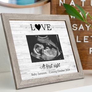 May include: A gray wood frame with a white background and a black heart with the word "LOVE" above it. The frame contains a black and white ultrasound image of a baby. The text "At first sight" is written below the image, with the baby's name and due date below that.