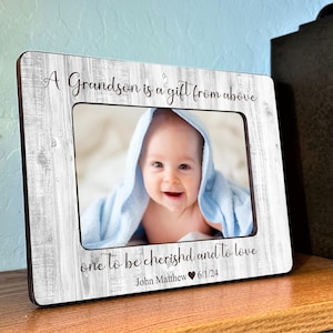 Puede incluir: Un marco de fotos de madera blanca con un borde negro y una foto de un beb&eacute; dentro. El marco tiene el texto "A Grandson is a gift from above one to be cherished and to love John Matthew 6/1/24" impreso en &eacute;l.