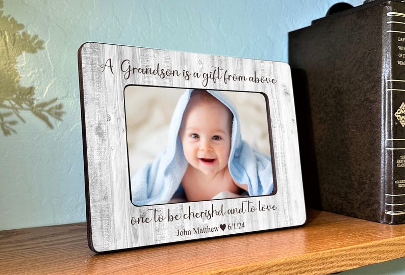 Puede incluir: Un marco de fotos de madera blanca con un borde negro y una foto de un beb&eacute; dentro. El marco tiene el texto "A Grandson is a gift from above one to be cherished and to love John Matthew 6/1/24" impreso en &eacute;l.
