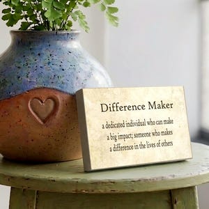 May include: A rectangular wooden sign with a gold-toned face featuring the black text: Difference Maker, a dedicated individual who can make a big impact; someone who makes a difference in the lives of others. The sign sits next to a ceramic vase.