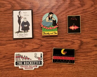 May include: A collection of enamel pins featuring various designs. One pin depicts Morticia and Uncle Fester from The Addams Family. Another shows Dorothy and Toto. Other pins include "A new day", The Rockettes, and the Andy Williams Moon River Theatre.