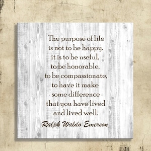 Peut inclure: Panneau carr&eacute; en bois avec un fond blanc effet grain de bois. Le panneau affiche une citation de Ralph Waldo Emerson en texte marron : "Le but de la vie n'est pas d'&ecirc;tre heureux..." Le panneau est mont&eacute; sur un mur beige.