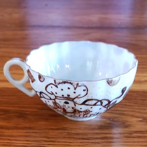 May include: A white teacup with a scalloped rim and a brown floral design. The cup has a handle and is made of porcelain.