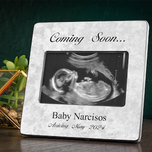May include: A white marble picture frame with a black border. The frame has a black and white ultrasound image inside and the text "Coming Soon... Baby Narcisos Arriving May 2024".
