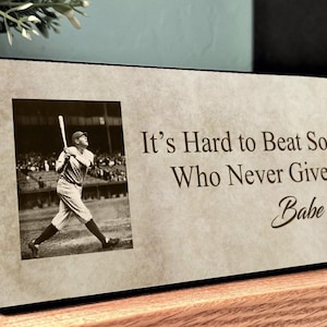 May include: A rectangular plaque with a sepia-toned image of Babe Ruth swinging a baseball bat. The plaque features the quote, "It's Hard to Beat Someone Who Never Gives Up" and Babe Ruth's signature. The plaque is displayed on a wooden shelf.