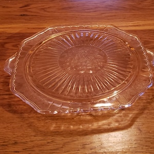 May include: A clear glass cake stand with a scalloped edge and a delicate, sunburst pattern. The stand is perfect for displaying cakes, pastries, or other desserts.