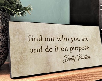 May include: A rustic wooden sign with a beige background and black lettering. The sign reads "find out who you are and do it on purpose" with the name "Dolly Parton" written in cursive below the quote.