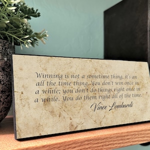 May include: A rectangular wooden sign with a vintage paper-like background. The sign features the quote "Winning is not a sometime thing..." by Vince Lombardi in black script. The sign is displayed on a wooden shelf with books and a plant.