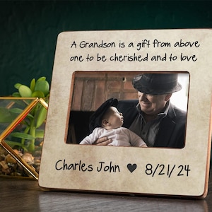 May include: A light brown wooden picture frame with a white mat. The frame has a quote that reads "A Grandson is a gift from above one to be cherished and to love". The frame also has the text "Charles John 8/21/24" and a heart symbol. The frame has a photo of a man holding a baby.