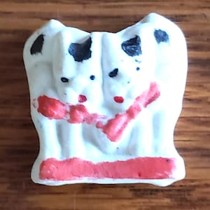 May include: A white ceramic figurine with two faces, each with black spots and red accents. The figurine has red lips and a red band at the base. The background is a wooden surface.