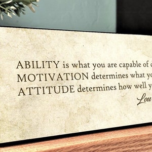 May include: A motivational quote by Lou Holtz on a wooden plaque with a vintage-style background. The quote reads: "ABILITY is what you are capable of doing. MOTIVATION determines what you do. ATTITUDE determines how well you do it."