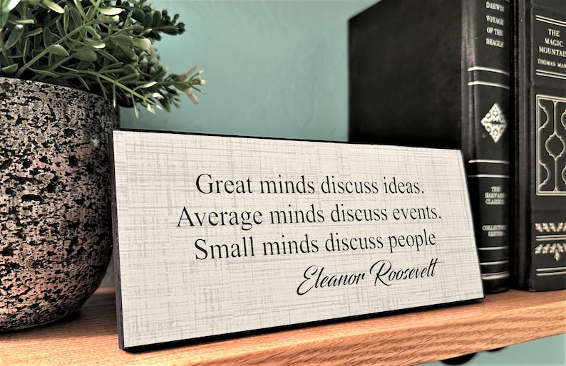 May include: A rectangular wooden sign with the quote "Great minds discuss ideas. Average minds discuss events. Small minds discuss people" by Eleanor Roosevelt. The sign has a light gray textured background and black text. The sign is on a wooden shelf.