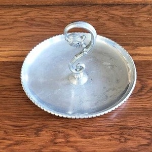 May include: Silver serving tray with a decorative handle in the shape of a stylized flower. The tray has a beaded edge and a smooth, reflective surface.