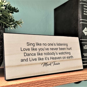 May include: A wooden sign with a black border, featuring a quote by Mark Twain: "Sing like no one's listening, Love like you've never been hurt, Dance like nobody's watching, and Live like it's Heaven on earth."