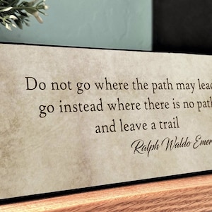 May include: A beige wooden plaque with a black border features a quote by Ralph Waldo Emerson: "Do not go where the path may lead, go instead where there is no path and leave a trail."