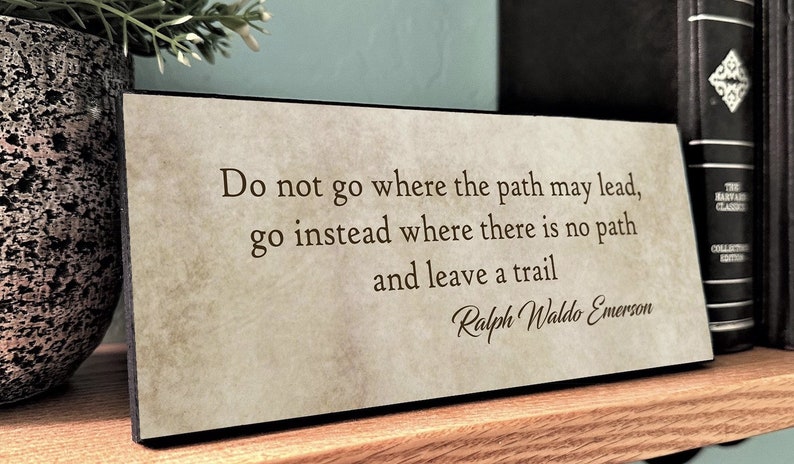 May include: A beige wooden plaque with a black border features a quote by Ralph Waldo Emerson: "Do not go where the path may lead, go instead where there is no path and leave a trail."
