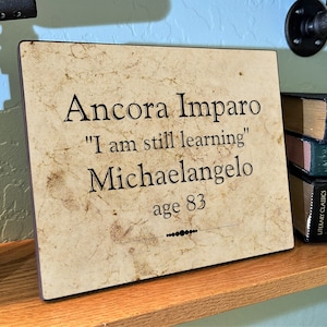 May include: A beige plaque with a brown border, featuring the quote "Ancora Imparo" in black lettering, which translates to "I am still learning." The quote is attributed to Michelangelo, with the age 83 listed below.
