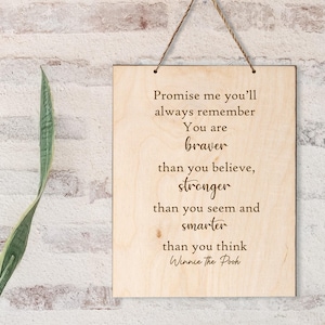 May include: Wooden sign with the quote "Promise me you'll always remember You are braver than you believe, stronger than you seem and smarter than you think Winnie the Pooh." The sign is hanging by a string.