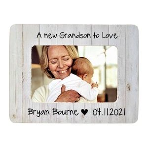 May include: A white wooden picture frame with a photo of a woman holding a baby. The frame has the text "A new Grandson to Love" at the top and "Bryan Bourne ♥ 04.11.2021" at the bottom.