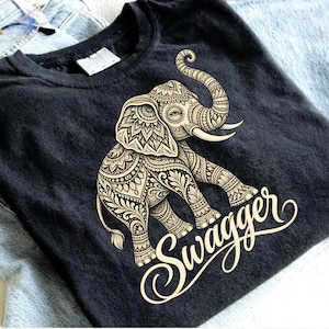 May include: A black t-shirt featuring a large, cream-colored graphic of an elephant decorated with intricate, repeating floral and geometric patterns. Below the elephant, the word "swagger" is printed in a flowing, cursive script font.