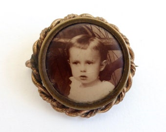 May include: A vintage gold-toned brooch with a round glass cameo featuring a black and white photo of a young boy. The cameo is surrounded by a rope-like design.