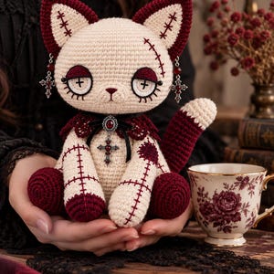 May include: A cream-coloured crocheted cat doll with dark red accents on the ears, paws, and tail. The doll features stitched detailing, cross-shaped eye pupils, and silver-toned cross charms hanging from the ears and a central red pendant.
