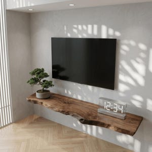 Könnte beinhalten: Ein schwebendes Wandregal aus Holz mit Naturkante an einer hellgrauen Wand unter einem Flachbildfernseher. Ein kleiner grüner Bonsai in einem grauen Topf und eine rechteckige Digitaluhr mit der Anzeige 12:34 stehen auf dem Regal.
