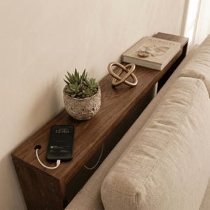 Puede incluir: Una mesa consola larga y estrecha de madera oscura situada detrás de un sofá beige. La superficie de la mesa sostiene una pequeña suculenta en una maceta de piedra texturizada, una escultura de nudo de metal dorado, un libro y un smartphone.