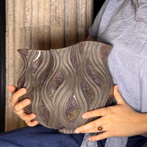 May include: A wide, textured ceramic vase with a dark grey, matte exterior featuring vertical, undulating wave patterns. The recessed areas between the waves reveal a shimmering, iridescent glaze with flecks of purple, gold, and bronze tones.