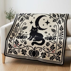 Puede incluir: Una manta de ganchillo color crema con un gato negro sentado bajo una luna creciente, rodeado de calabazas y hojas de roble. El diseño incluye pequeñas bayas rojas y un borde decorativo con un patrón gráfico detallado.