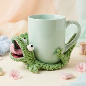 May include: A light green ceramic mug held by a green crocheted frog cozy. The frog features a wide open mouth with a pink tongue, two white eyes with black pupils, and small limbs that wrap around the base and sides of the mug.