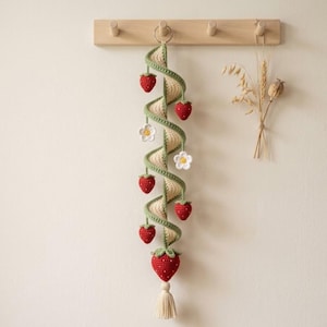 May include: A spiral crochet wall hanging featuring a cream-coloured centre with a green vine winding around it. The piece is decorated with several small red strawberries with green tops and two white flowers with yellow centres, ending in a tassel.