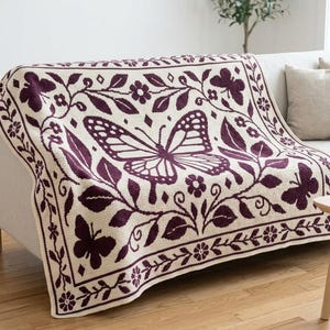 May include: A cream-colored knit throw blanket featuring a dark purple pattern of a large central butterfly surrounded by smaller butterflies, leaves, and flowers, all enclosed within a decorative border of repeating floral motifs.