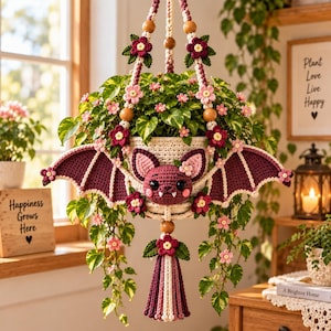 May include: A cream-colored crocheted hanging plant holder featuring a central bat face with large ears, fangs, and dark purple wings. The holder is decorated with small pink and dark purple crocheted flowers, wooden beads, and a long tassel.