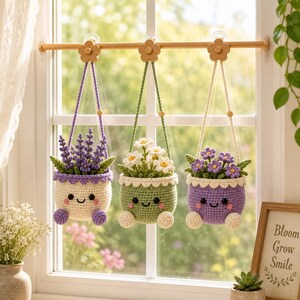 Puede incluir: Tres macetas colgantes de ganchillo con caras sonrientes, cada una con un arreglo floral diferente. Las macetas cuelgan de una barra mediante ganchos de madera con forma de flor. Las flores incluyen lavanda morada, margaritas blancas y flores moradas.