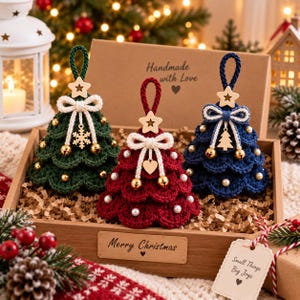 Può includere: Tre decorazioni per albero di Natale all'uncinetto in verde scuro, rosso intenso e blu navy, ciascuna sormontata da una stella in legno, un fiocco bianco e piccoli campanellini dorati. Sono esposte in una scatola con la scritta Merry Christmas.