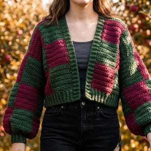 Può includere: Un cardigan corto all'uncinetto con motivo a scacchi in verde scuro e bordeaux intenso. Il capo presenta maniche lunghe e voluminose con polsini a coste, uno scollo a V e un'apertura frontale senza bottoni, indossato sopra un top grigio scuro.