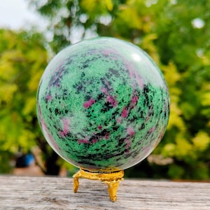 May include: A polished spherical stone featuring a mottled pattern of vibrant green, deep magenta, and black mineral inclusions. The sphere rests securely upon a small, textured gold-colored metal tripod stand with three distinct feet.