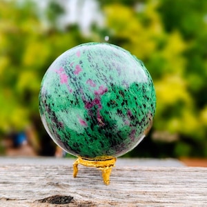 Peut inclure: Une pierre sphérique polie présentant une base vert vif avec des inclusions rose magenta dispersées et des stries minérales noires foncées, reposant solidement sur un petit support de présentation métallique doré à trois pieds.