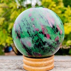 Puede incluir: Una piedra esférica pulida que presenta una base de color verde vibrante con inclusiones de color rosa magenta dispersas y vetas minerales de color negro oscuro, apoyada sobre un soporte de madera clara circular con diseño escalonado.